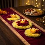 Flower rangoli diya set styled along a maroon and gold table runner with a mithai thali and fairy lights for a Diwali dinner at home.