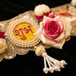 Close-up of Shubh Labh medallion embroidered in red on gold, surrounded by ivory pearls, with crystal-accented fabric rose and scalloped gold lace border